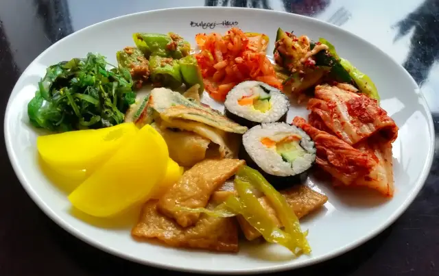Ovaler Bulgogi-Haus-Teller mit gemischten Banchan: Seetangsalat, gefüllte Peperoni, Gemüsepfannkuchen, Gimbap-Röllchen, Kimchi, Gurkensalat, eingelegter gelber Rettich und gebratener Fischkuchen.