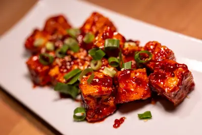Knusprig gebratene Tofuwürfel in roter Gochujang-Sauce, bestreut mit Frühlingszwiebeln und Sesam, auf weisser Platte.