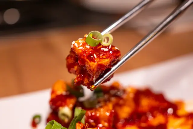 Metallstäbchen halten ein knusprig frittiertes Hähnchenstück in glänzender roter Yangnyeom-Sauce mit Sesam und einem Frühlingszwiebelring.