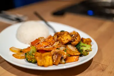 Teller mit würzig gebratenem Gemüse, Tofu, Brokkoli, Karotten und Champignons mit Sesam, dazu ein Häufchen weißer Reis.