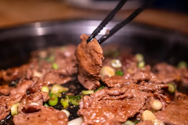 Eine Grillzange hebt ein Stück mariniertes Rindfleisch vom Tischgrill, darunter brutzelt Bulgogi mit Frühlingszwiebeln.