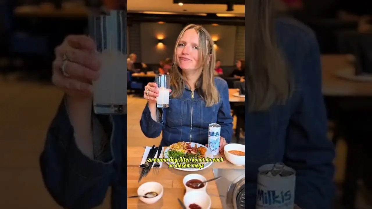 Eine lächelnde Frau sitzt in einem Restaurant an einem Esstisch, hält ein Glas mit einem milchigen Getränk hoch und stellt eine Dose Milkis sowie Teller mit Essen vor.